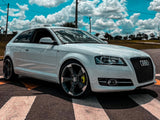 A white Audi car is parked diagonally on a road's crosswalk, under a partly cloudy blue sky. Power lines and grassy fields are visible in the background.