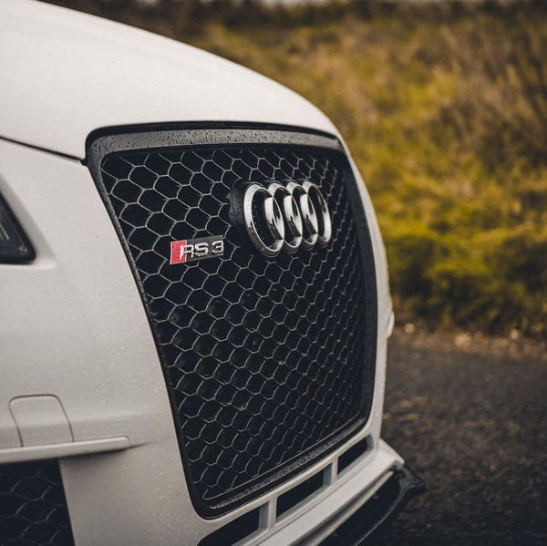 A car's front grille displays the Audi logo and "RS3" badge, set against a hexagonal mesh design, with grass visible in the background.
