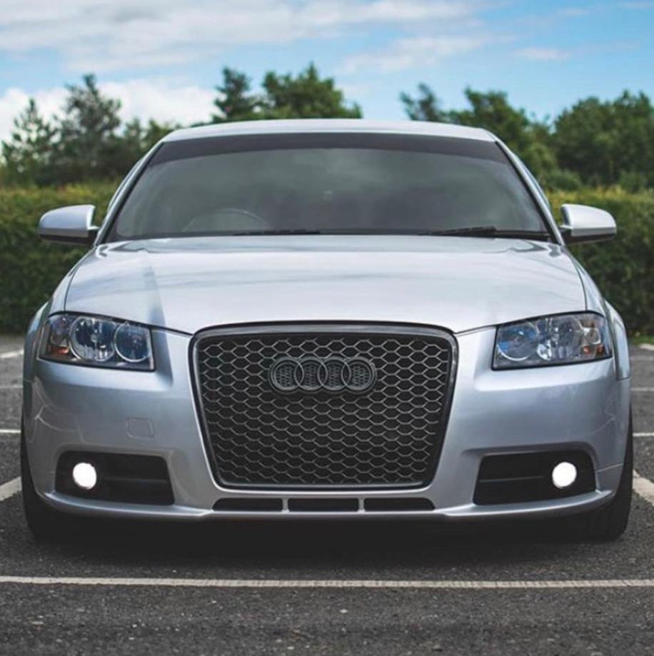 A silver car with a distinctive honeycomb grille is parked, surrounded by greenery and a partly cloudy sky. The car features circular headlights and the Audi logo on its front.