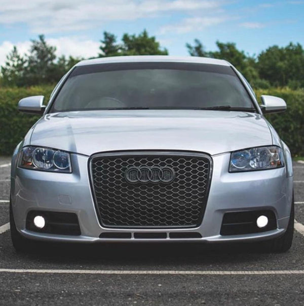 A silver car with a distinctive honeycomb grille is parked, surrounded by greenery and a partly cloudy sky. The car features circular headlights and the Audi logo on its front.