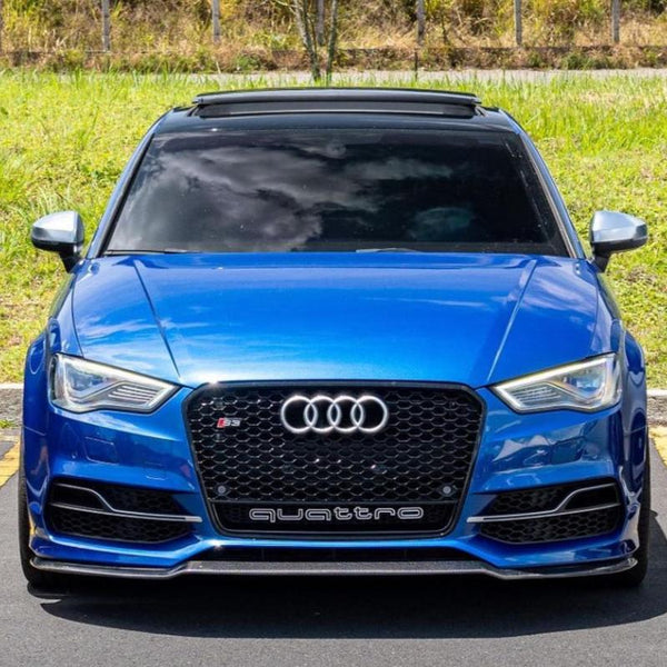 A blue Audi S3 with a sunroof and "quattro" on the grille is parked on asphalt, surrounded by grass and trees.