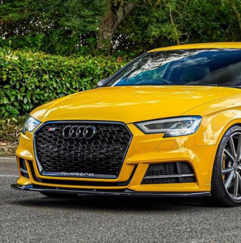 A yellow sports car is parked on a road, featuring a black grille and the Audi logo, with "quattro" text visible, surrounded by lush green foliage.
