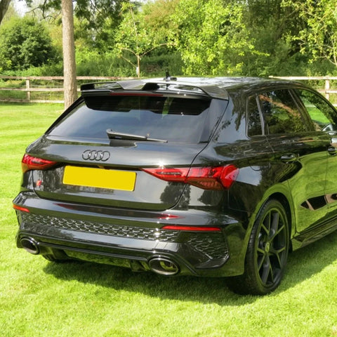 A black Audi car is parked on a grassy area surrounded by trees and a wooden fence. It has prominent dual exhaust pipes and tinted windows.