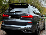 A dark gray BMW X7 parked on a leaf-covered path, surrounded by autumn trees. The car's rear features red taillights and dual exhaust. Visible text includes 