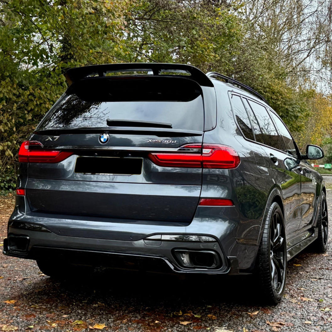 A dark gray BMW X7 parked on a leaf-covered path, surrounded by autumn trees. The car's rear features red taillights and dual exhaust. Visible text includes "X7" and "Xdrive30d."