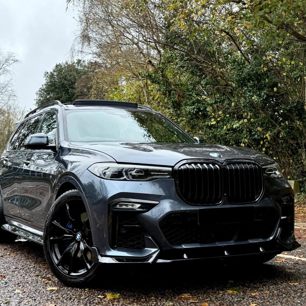 A gray SUV is parked on a gravel path, surrounded by lush green trees and overcast skies, showcasing its sleek front design and black accents.