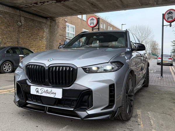 A silver BMW SUV is parked under a bridge with "A Detailing" on its front plate. Two "no cars allowed" signs are visible, surrounded by brick buildings and parked vehicles.