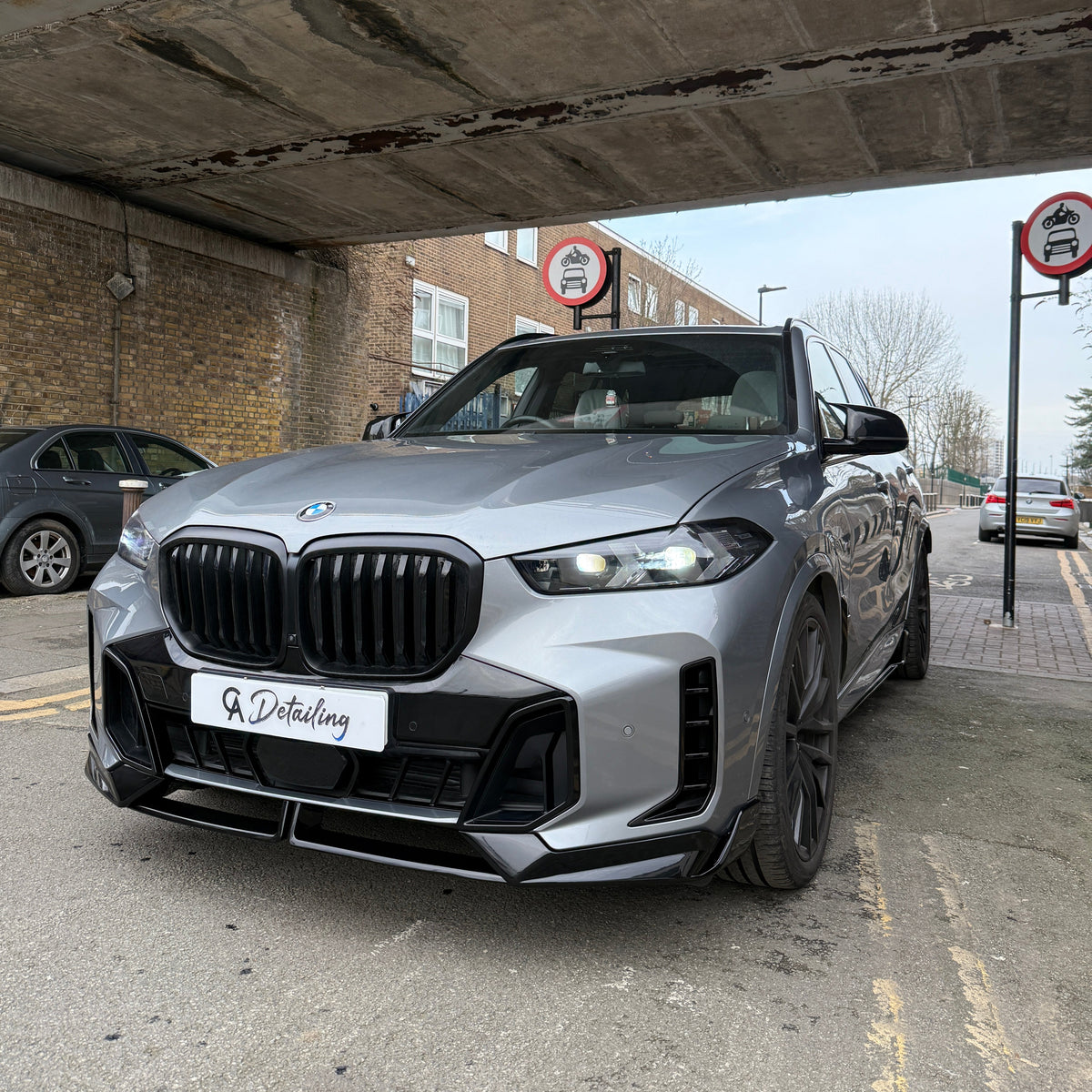 A silver BMW SUV is parked under a bridge with "A Detailing" on its front plate. Two "no cars allowed" signs are visible, surrounded by brick buildings and parked vehicles.