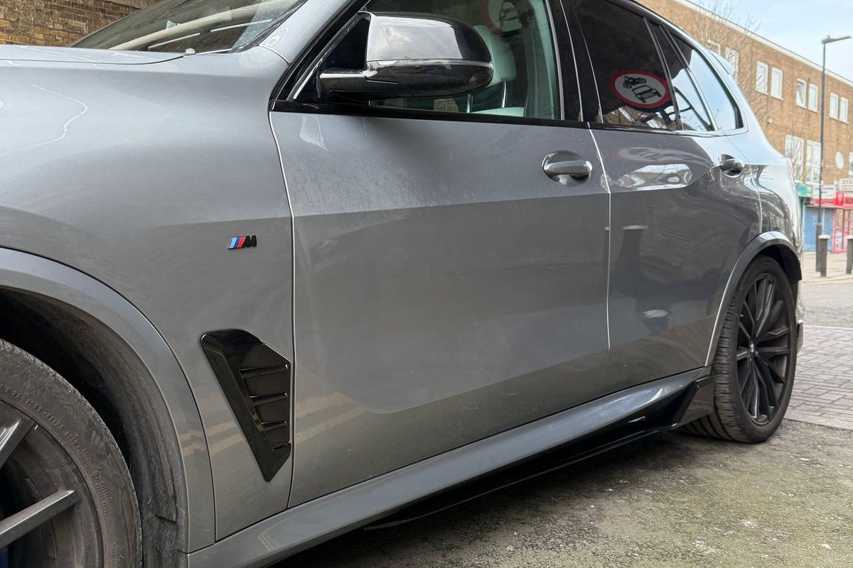 A gray SUV is parked under a concrete structure beside a building. It features a black M badge on the side, indicating its model variant.