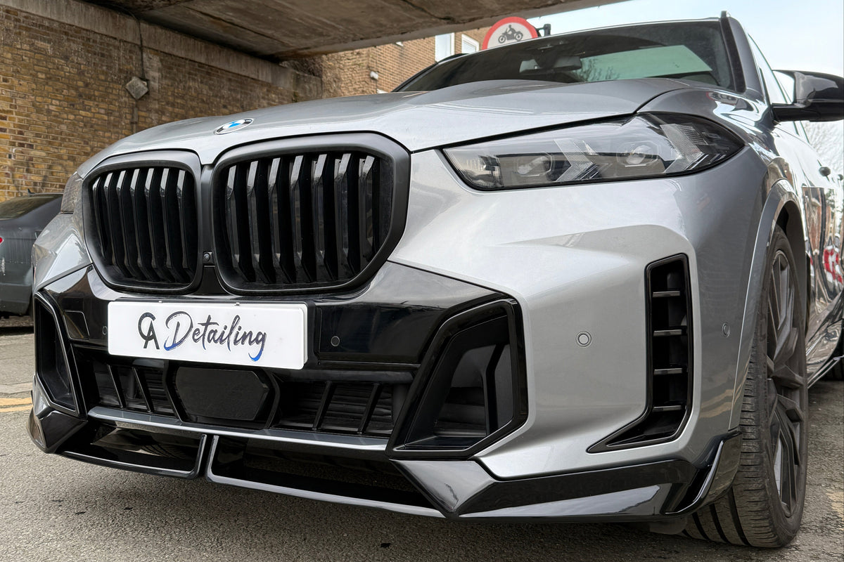 A silver BMW with a prominent grille is parked under an urban bridge. A sign reads "A Detailing." The setting includes brick walls and street signs.