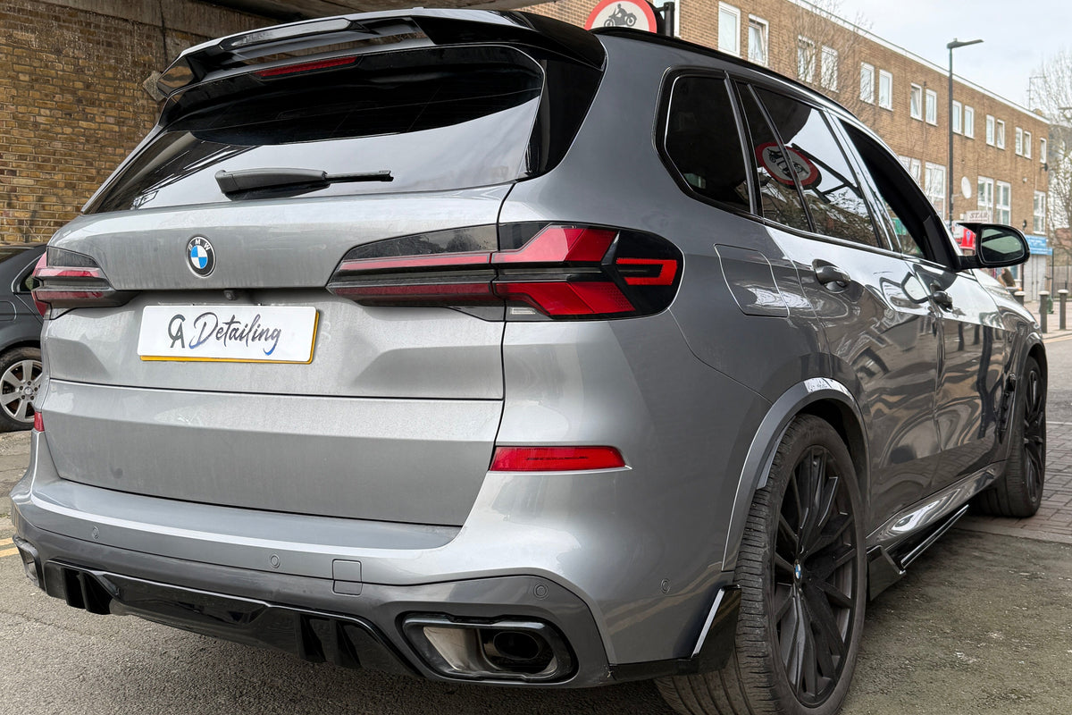 A silver BMW SUV is parked under a bridge, displaying "A Detailing" on the license plate. Nearby, brick buildings and street signs are visible in an urban setting.