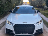 A white Audi car is parked on a narrow stone path, flanked by lush trees. The vehicle's front features the Audi logo, with 