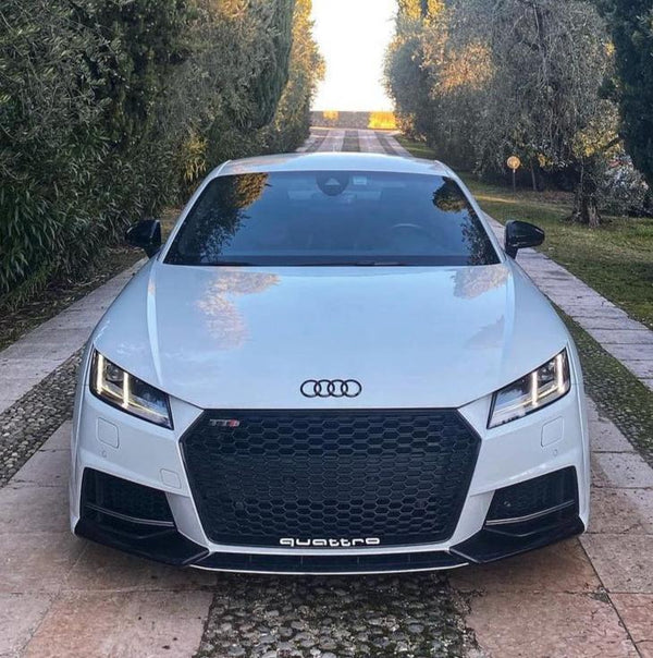 A white Audi car is parked on a narrow stone path, flanked by lush trees. The vehicle's front features the Audi logo, with "TTS" and "quattro" displayed.