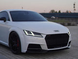 A white Audi sports car is parked, displaying sleek black rims and LED headlights, against a backdrop of tiles and a distant urban skyline. 