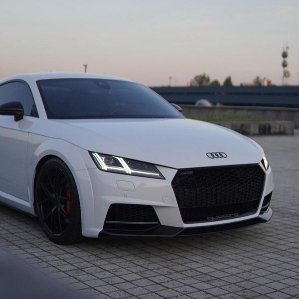 A white Audi sports car is parked, displaying sleek black rims and LED headlights, against a backdrop of tiles and a distant urban skyline. "quattro" is visible on the grille.