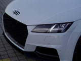 A white Audi car is parked on a paved surface. The front grille displays the Audi logo. 