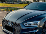 A sleek black Audi car is parked, with its distinctive grille and emblem visible. It rests on a paved area beside a grassy field and trees. The text reads 