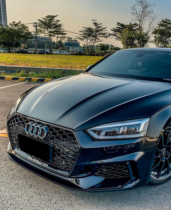 A sleek black Audi car is parked, with its distinctive grille and emblem visible. It rests on a paved area beside a grassy field and trees. The text reads "quattro".