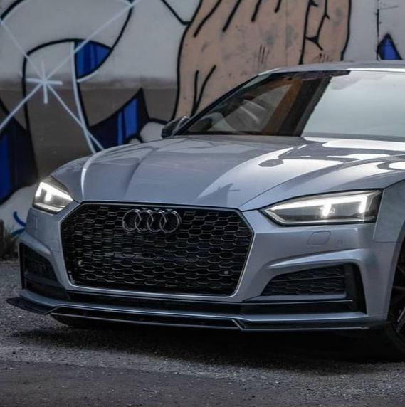 A silver car with a sleek design is parked on a gravel surface, its headlights on. It stands in front of a colorful graffiti mural, featuring abstract shapes and patterns.