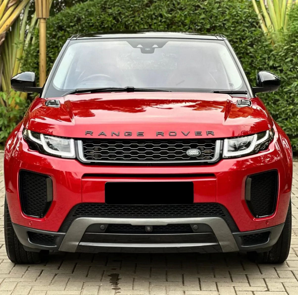 A red Range Rover is parked on a brick driveway, surrounded by lush greenery. The front of the car prominently displays the "RANGE ROVER" and "LAND ROVER" logos.
