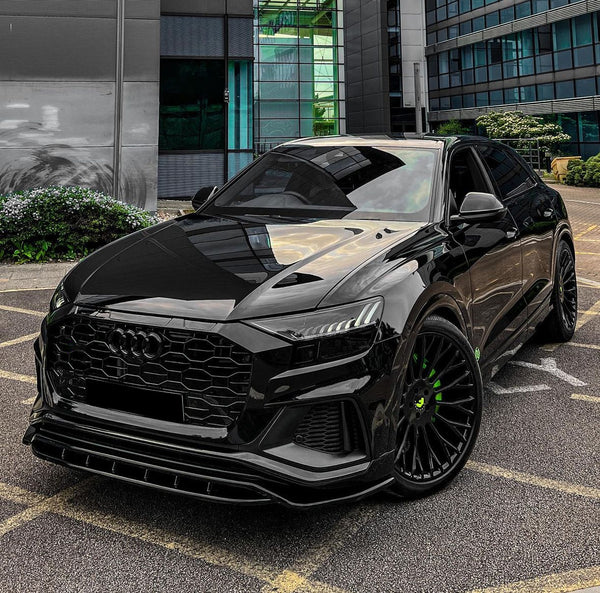 A sleek black car is parked on a lot, featuring green-accented wheels, in front of a modern glass and concrete building.