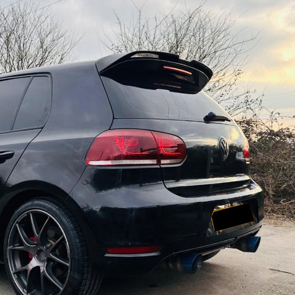 A black car is parked on a concrete surface, featuring prominent red taillights and tinted windows. Bare trees and a cloudy sky create a wintery backdrop. License plate is obscured.