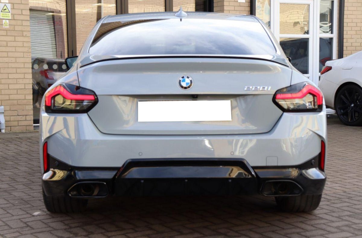 A gray BMW 220i is parked on a brick driveway. The car's rear view features sleek taillights and a visible badge. A building with glass doors and windows surrounds it.