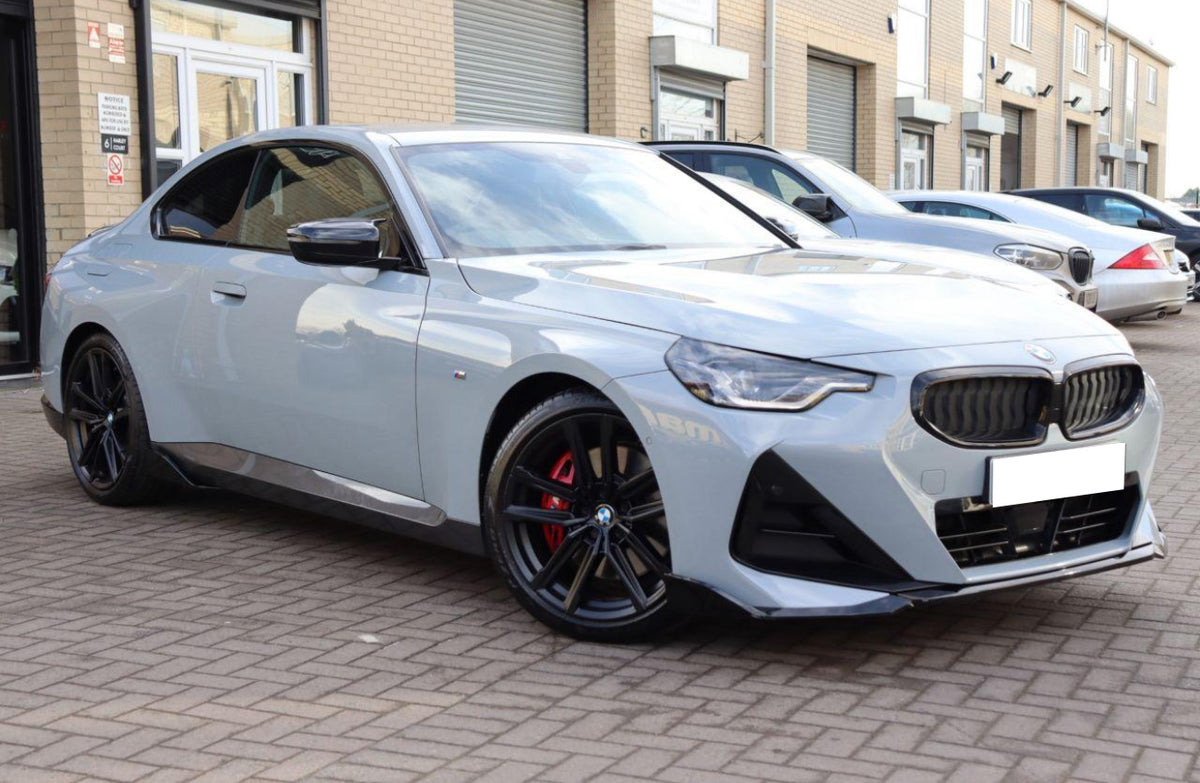 A silver car is parked on a brick driveway, its sleek design complemented by black wheels and red brake calipers. Surrounding it are more vehicles and a building with roller doors.