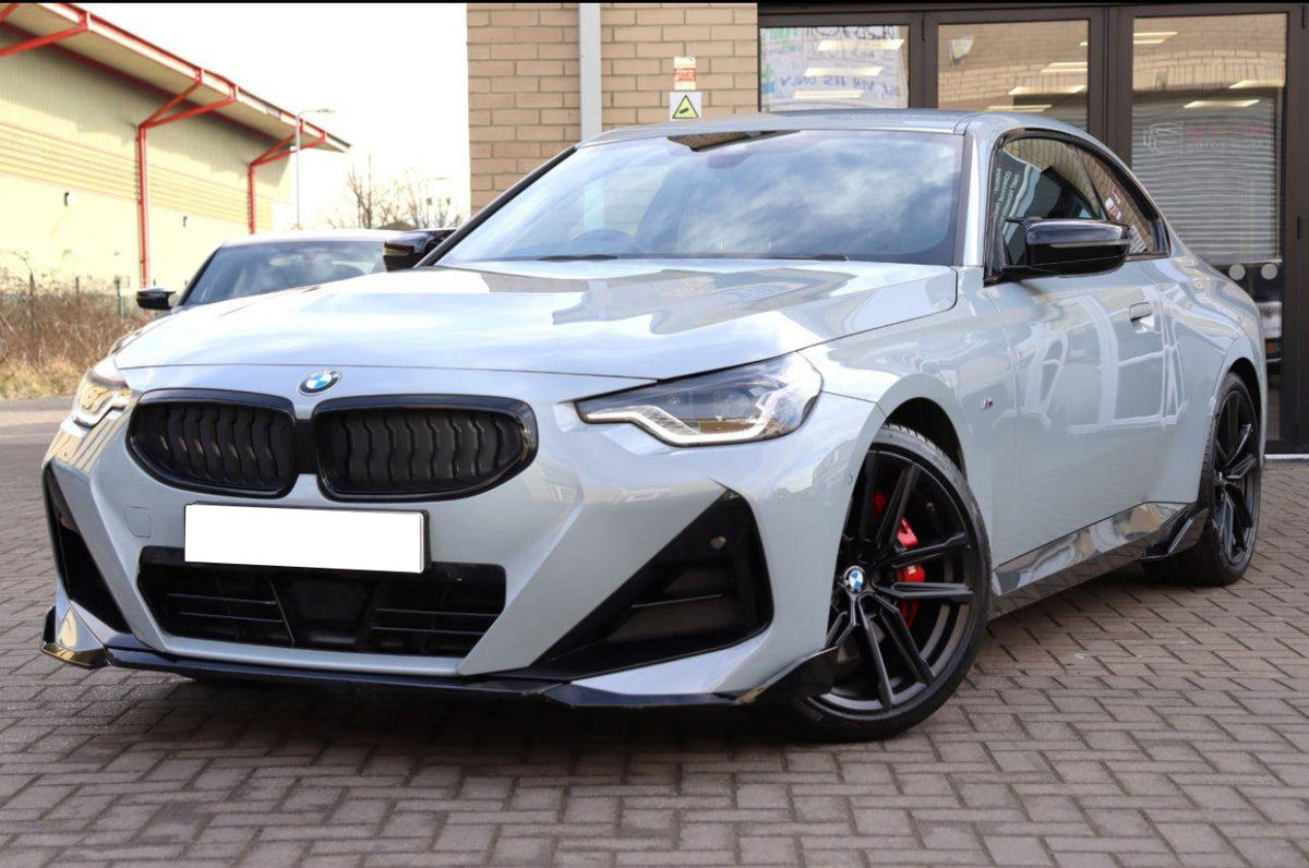 A silver BMW car is stationary on a brick driveway, facing forward, with a modern building in the background. The car has sleek black rims and red brake calipers.