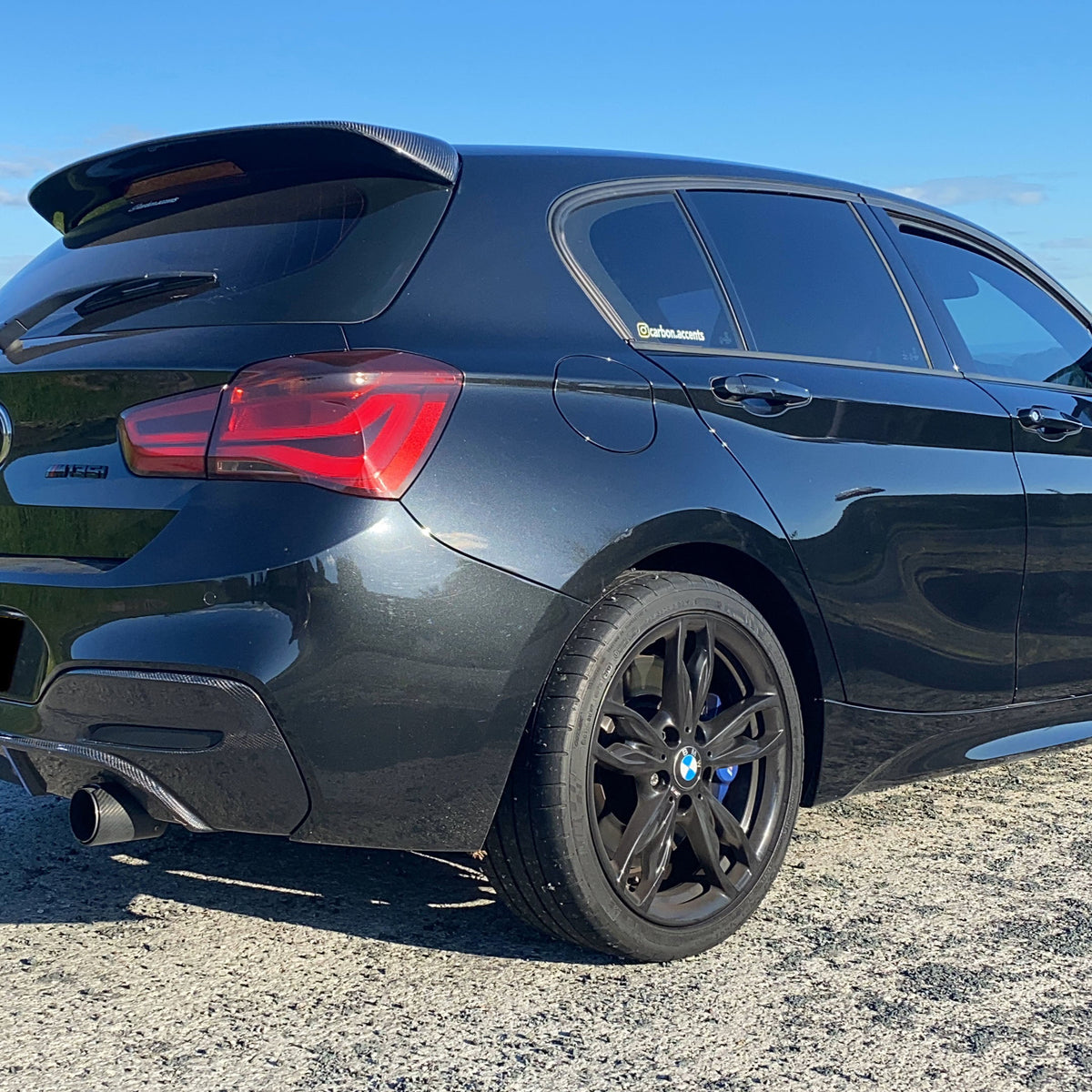 Car parked on gravel, with a shiny black exterior and dark alloy wheels. Visible text on window: "@carbon.accenta". Bright blue sky forms the background.