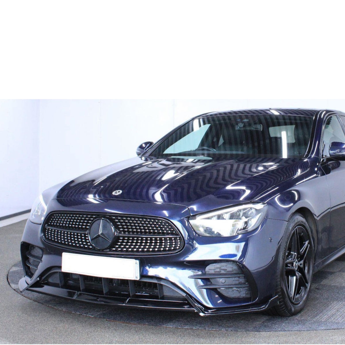 A sleek, dark blue Mercedes-Benz car is parked in a well-lit indoor space, featuring a prominent front grille and modern headlights, with reflective surfaces highlighting its polished finish.