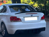 A white BMW car is parked, facing away, showing taillights, on a paved surface surrounded by trees and greenery. A parking sign is visible in the background.