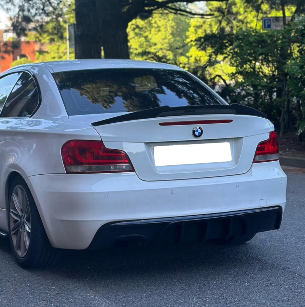A white BMW car is parked, facing away, showing taillights, on a paved surface surrounded by trees and greenery. A parking sign is visible in the background.