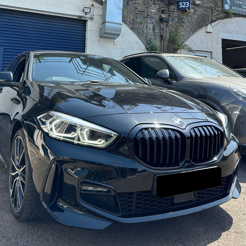 A sleek black BMW parked on a sunny day, next to another car, in front of a brick building with a blue garage door. Signs read "523" and "CAR KEY...AUTO ELECTRICS".
