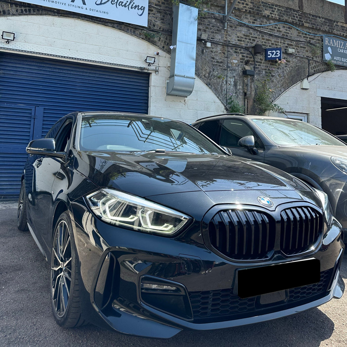 A black car with distinctive headlights is parked in front of a building featuring blue shutters and a sign reading "Automotive Styling & Detailing." A second vehicle is parked nearby.