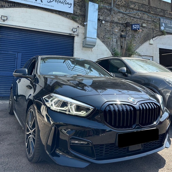 A black car with distinctive headlights is parked in front of a building featuring blue shutters and a sign reading "Automotive Styling & Detailing." A second vehicle is parked nearby.