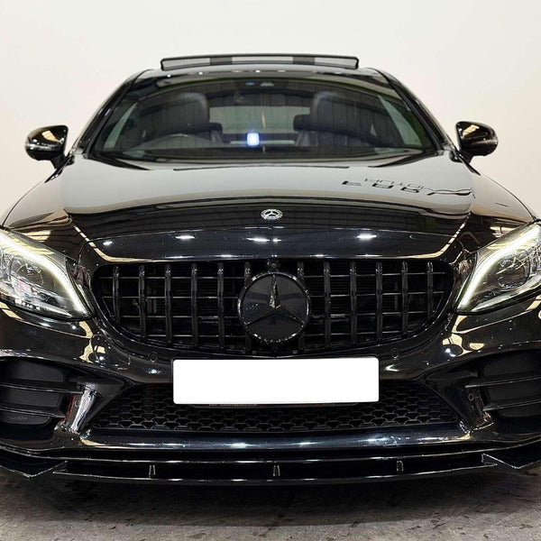 A black luxury sedan with illuminated headlights is parked on a concrete floor in a white-walled room. The license plate is blank, and a Mercedes-Benz emblem is visible.