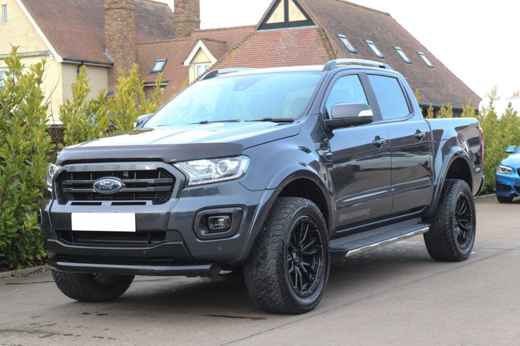 A gray Ford Ranger Wildtrak pickup truck is parked on a driveway. The background features residential houses with sloped roofs and green shrubs lining the pathway.