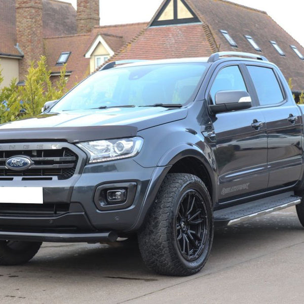 A gray Ford Ranger Wildtrak pickup truck is parked on a driveway. The background features residential houses with sloped roofs and green shrubs lining the pathway.