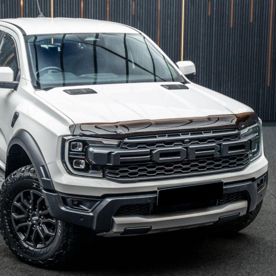 Pickup truck with "FORD" on the grille, parked in a covered area with vertical wood slat walls. White body and large tires convey a robust, contemporary design.
