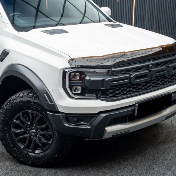 White pickup truck with black grille showing large "FORD" letters, parked indoors on a carpeted surface, surrounded by a dark paneled background.