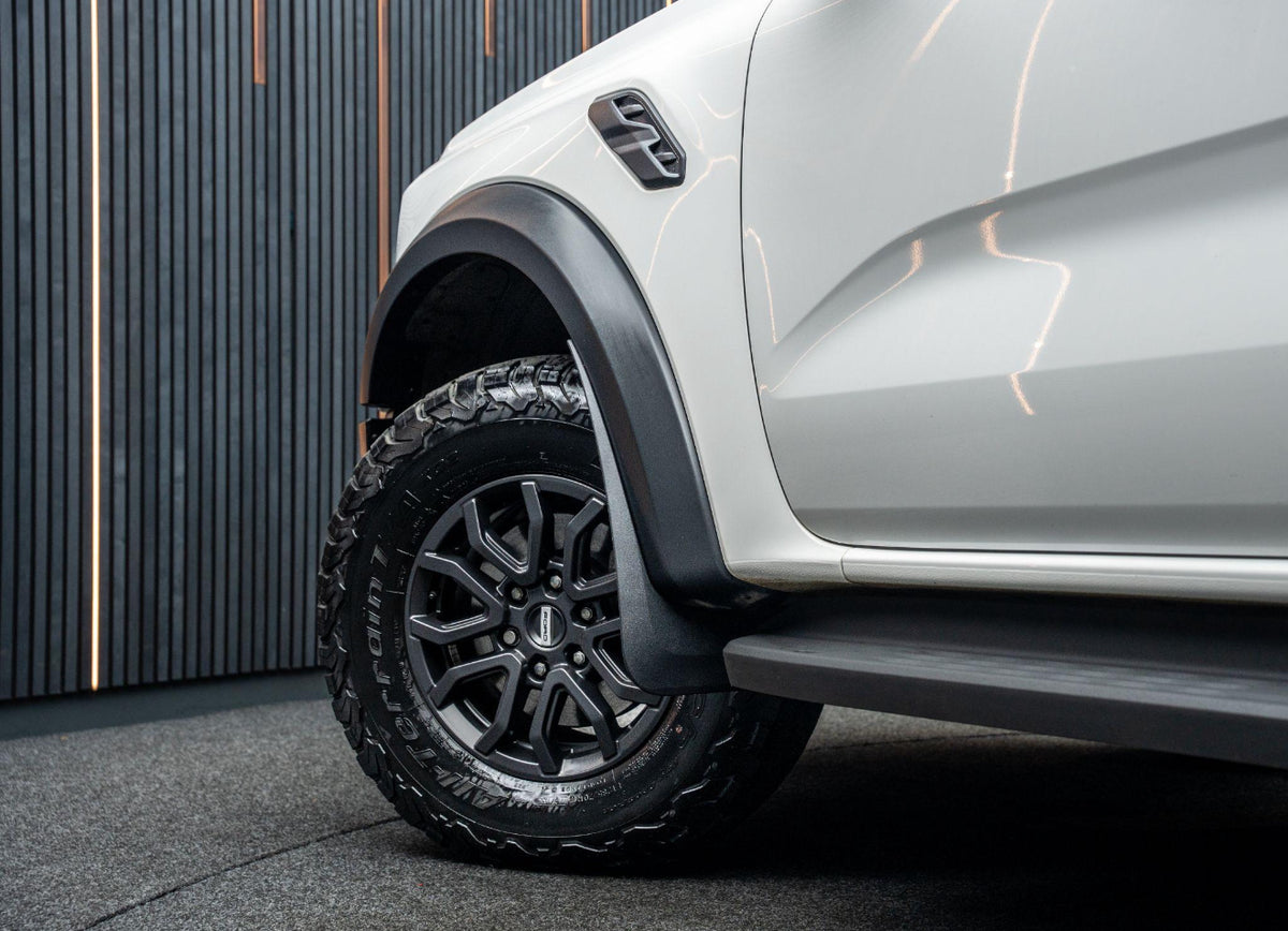 A rugged car tire rests on a black wheel, with visible tread and "Terrain T/A" text. The white vehicle body stands in a dimly lit room with vertical striped walls.