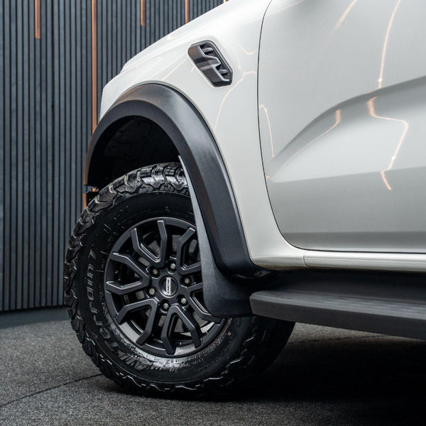 A rugged car tire rests on a black wheel, with visible tread and "Terrain T/A" text. The white vehicle body stands in a dimly lit room with vertical striped walls.