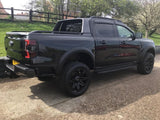 A black pickup truck is parked on a paved area, surrounded by green grass and trees. The vehicle has tinted windows and large, rugged tires, with reflections visible on its body.