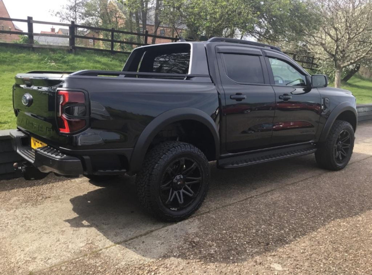 A black pickup truck is parked on a paved area, surrounded by green grass and trees. The vehicle has tinted windows and large, rugged tires, with reflections visible on its body.