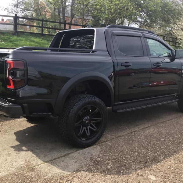 A black pickup truck is parked on a paved area, surrounded by green grass and trees. The vehicle has tinted windows and large, rugged tires, with reflections visible on its body.