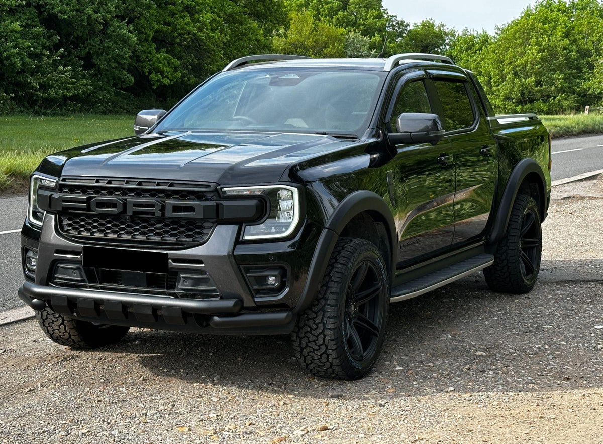 A black pickup truck is parked on a gravelly area by a roadside, surrounded by lush green trees. The grille prominently displays "FORD" in bold lettering.
