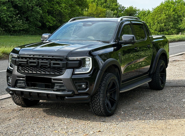 A black pickup truck is parked on a gravelly area by a roadside, surrounded by lush green trees. The grille prominently displays "FORD" in bold lettering.