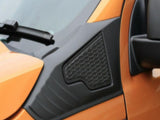 An orange vehicle with a black plastic panel featuring a hexagonal pattern, located near the side mirror and windshield wiper, set against a smooth, light background.