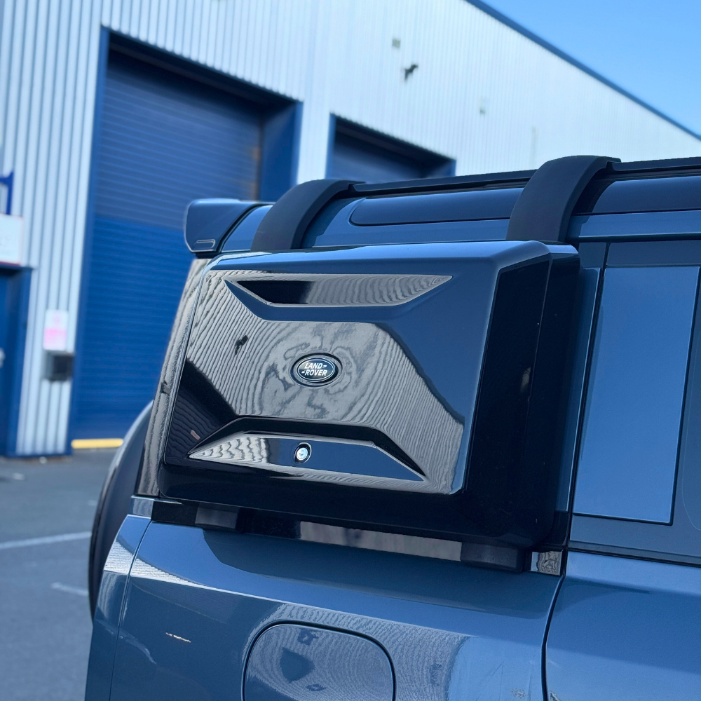 A Land Rover spare tire case is mounted on the back of a vehicle. It features the Land Rover logo, set outdoors near industrial blue garage doors.